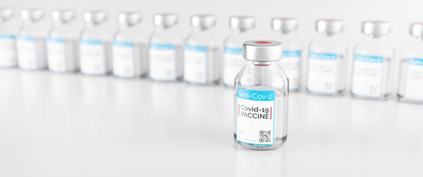 Ein Glasfl�schchen gef�llt mit heller Fl�ssigkeit (Impfstoff) und der Aufschrift "Sars-Cov-2 - Covid-19 VACCINE". Im Hintergrund viele weitere solcher Glasfl�schchen nebeneinander aufgereiht.