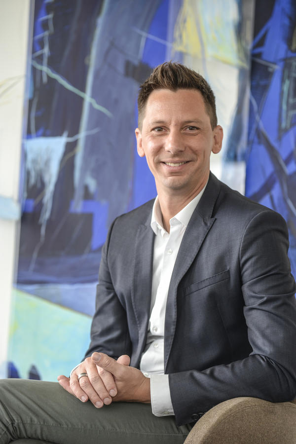 Landrat Andreas M�ller in wei�em Hemd, dunkelblauem Sakko und dunkelgr�ner Hose sitzt auf einem beige bezogenen Stuhl vor einem blau, wei�, gelben Gem�lde.