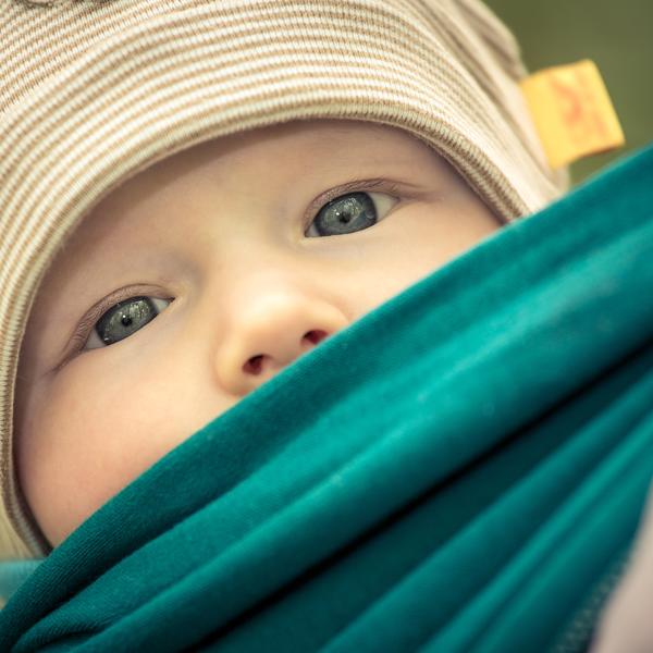 Ein kleines Kind mit einer wei�-gelb-gestreiften M�tze schaut aus einer dunkelgr�nen Trage heraus. Es sind nur dessen glas-blauen Augen und die kleine Nase zu sehen.